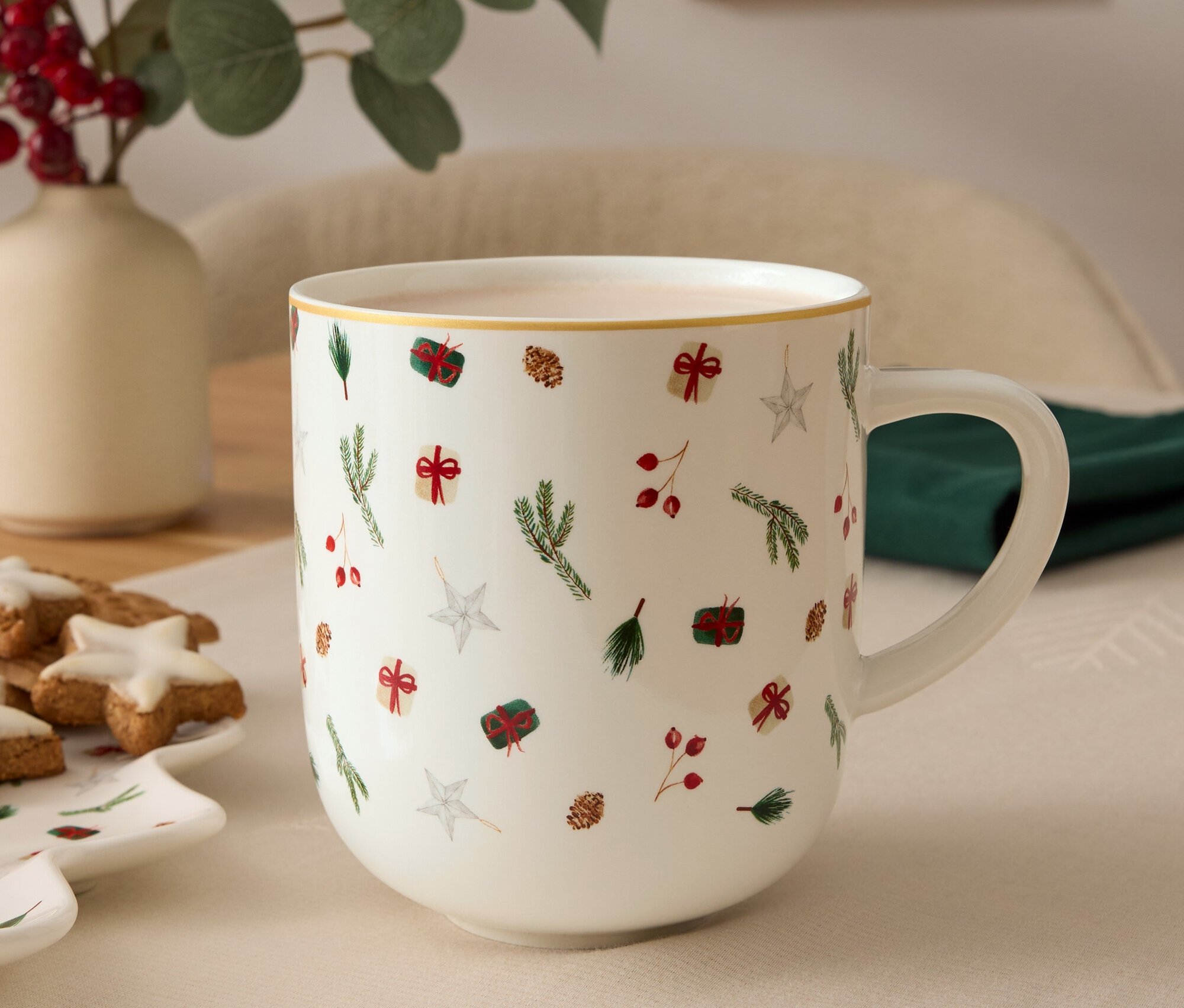 Im Vordergrund befindet sich eine Tasse mit Weihnachtsmotiven, im Hintergrund eine Vase mit Pflanzen und ein Teller mit Keksen.
