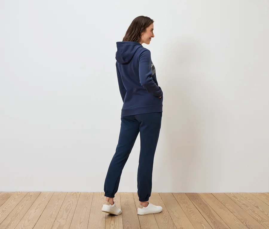 Eine Frau in marineblauem Hoodie und Trainingshose steht seitlich auf einem Holzboden.