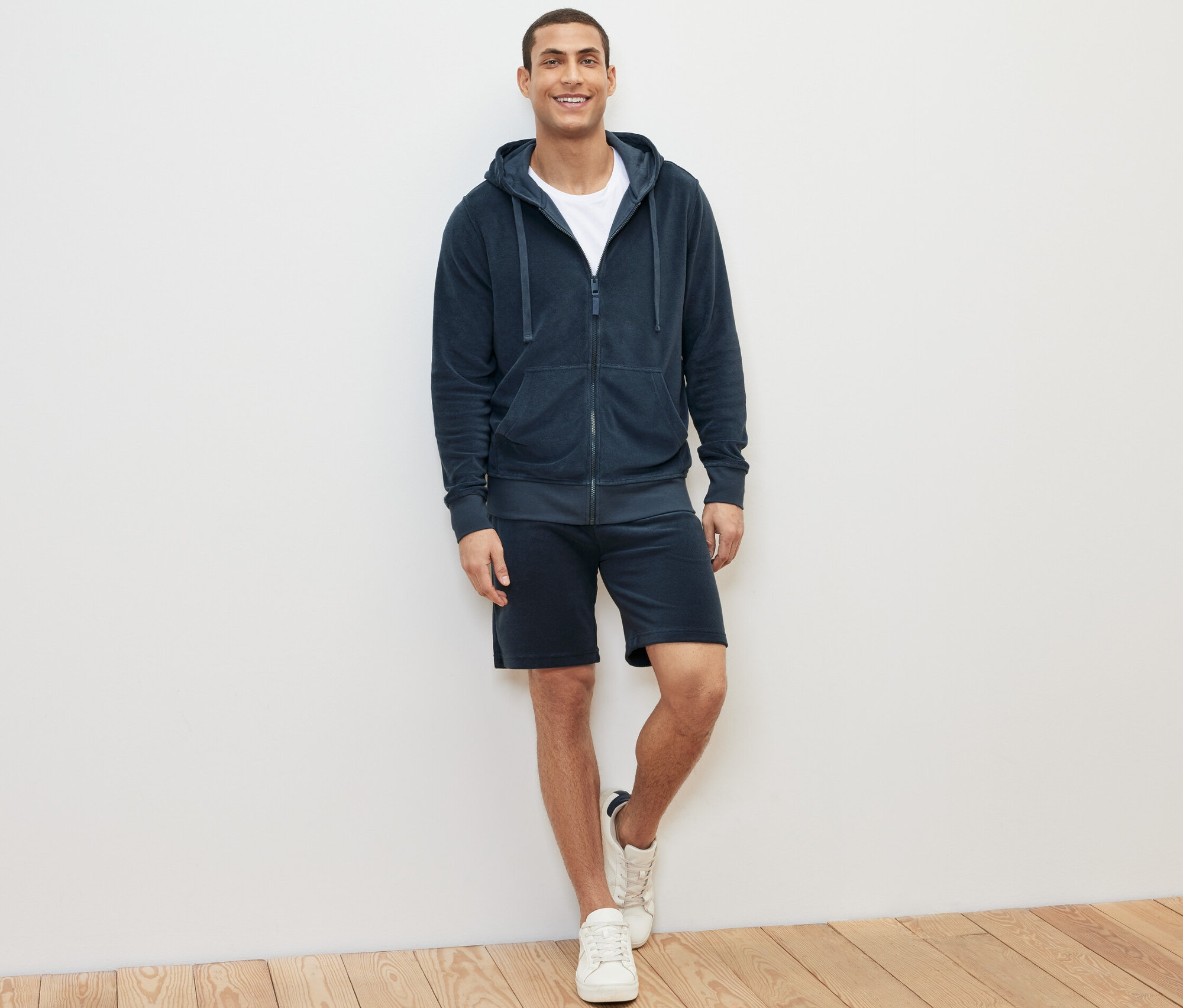 Mann steht und trägt eine blaue Frottee-Kapuzenjacke, T-Shirt und Shorts.