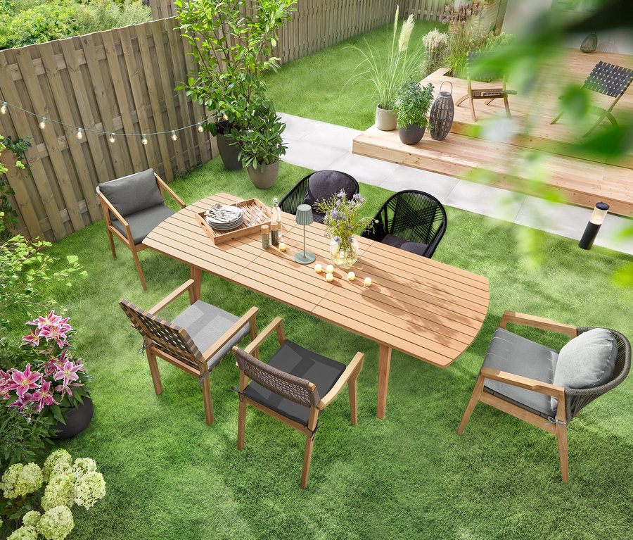 Ein ausziehbarer Gartentisch mit abgerundeten Fronten steht auf einer grünen Rasenfläche, umgeben von mehreren Gartenstühlen. Eine Solar-Lichterkette »Glühbirnen« hängt über dem Tisch, und eine LED-Akku-Outdoor-Tischleuchte, salbei, steht darauf. Mehrere 6 LED-Outdoor-Akku-Teelichter sind auf dem Tisch verteilt.