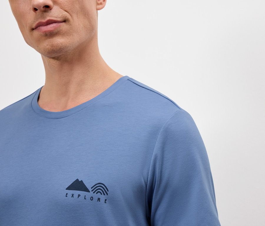 Mann trägt ein blaues T-Shirt mit dem Aufdruck "Explore", der aus einem Bergsymbol und Regenbogen besteht.