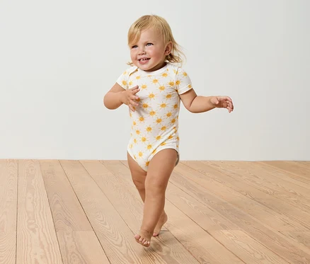 Ein blondes Kleinkind in einem Body mit Sonnenmuster geht auf einem Holzboden.