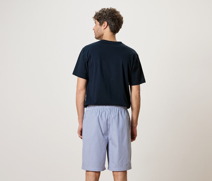 Ein Mann von hinten, trägt ein marineblaues T-Shirt und blau-weiss gestreifte Shorts.