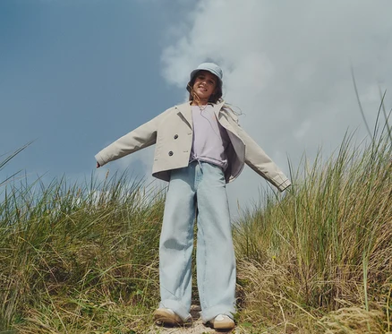 Ein junges Mädchen steht in einem Feld mit hohem Gras, trägt eine helle Jacke, weite Jeans und einen passenden Hut.