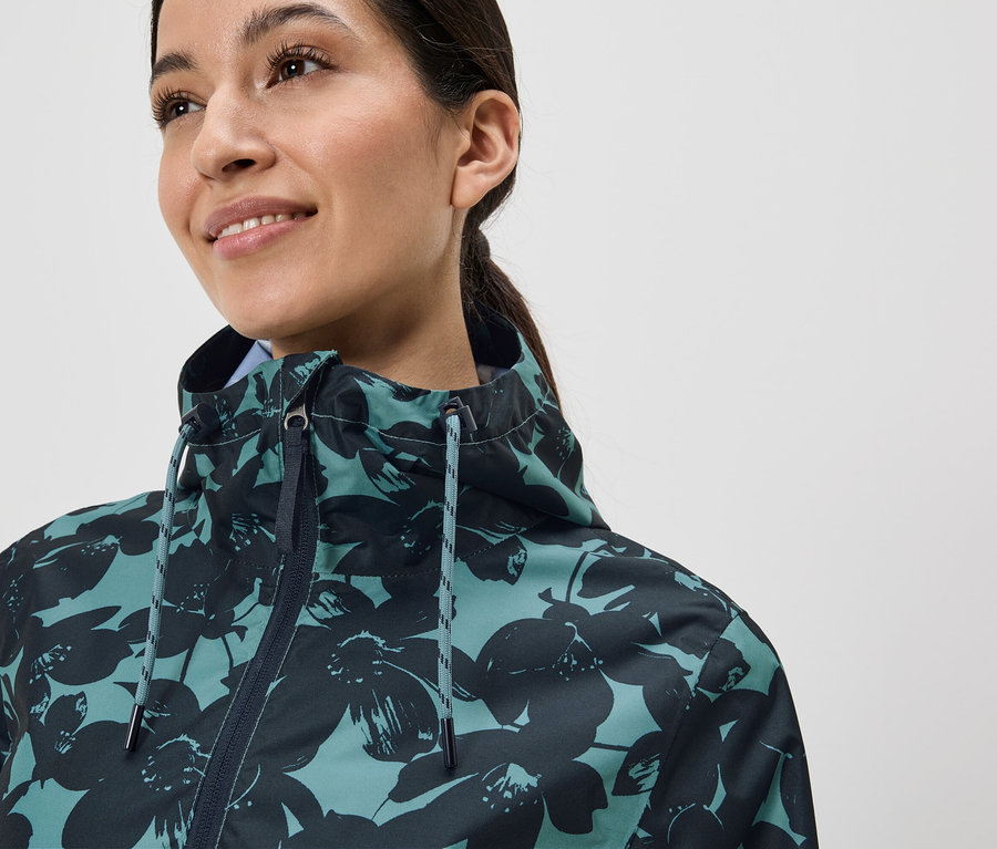 Lächelnde Frau mit Pferdeschwanz trägt eine blaugrüne Jacke mit dunklen Blumen.