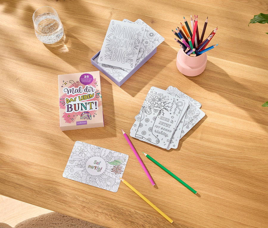 Auf einem Tisch liegen Malbücher, Karten mit Motiven zum Ausmalen, Buntstifte in einem rosa Stifthalter und ein Glas Wasser.