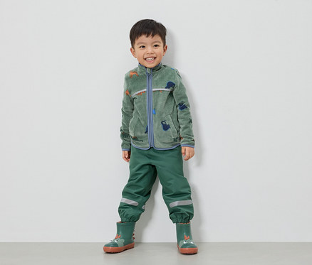 Ein lächelnder Junge steht in grüner Fleecejacke mit Dinosauriern, grüner Hose und grünen Gummistiefeln vor einer weißen Wand.