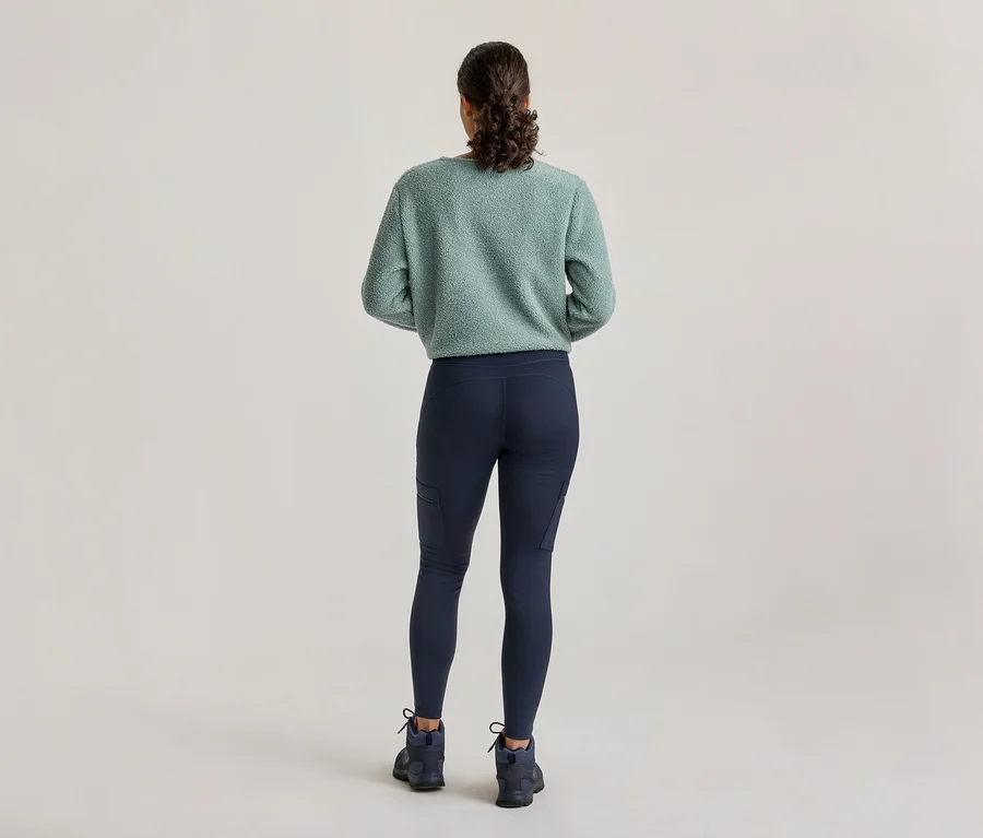 Eine Person von hinten, die einen hellgrünen flauschigen Pullover, dunkelblaue Leggings mit Seitentaschen und dunkelblaue Wanderschuhe trägt.