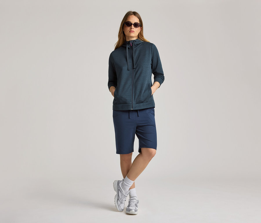 Frau mit Sonnenbrille trägt marineblaue Kapuzenjacke und Shorts. Die Kapuzenjacke ist offen.