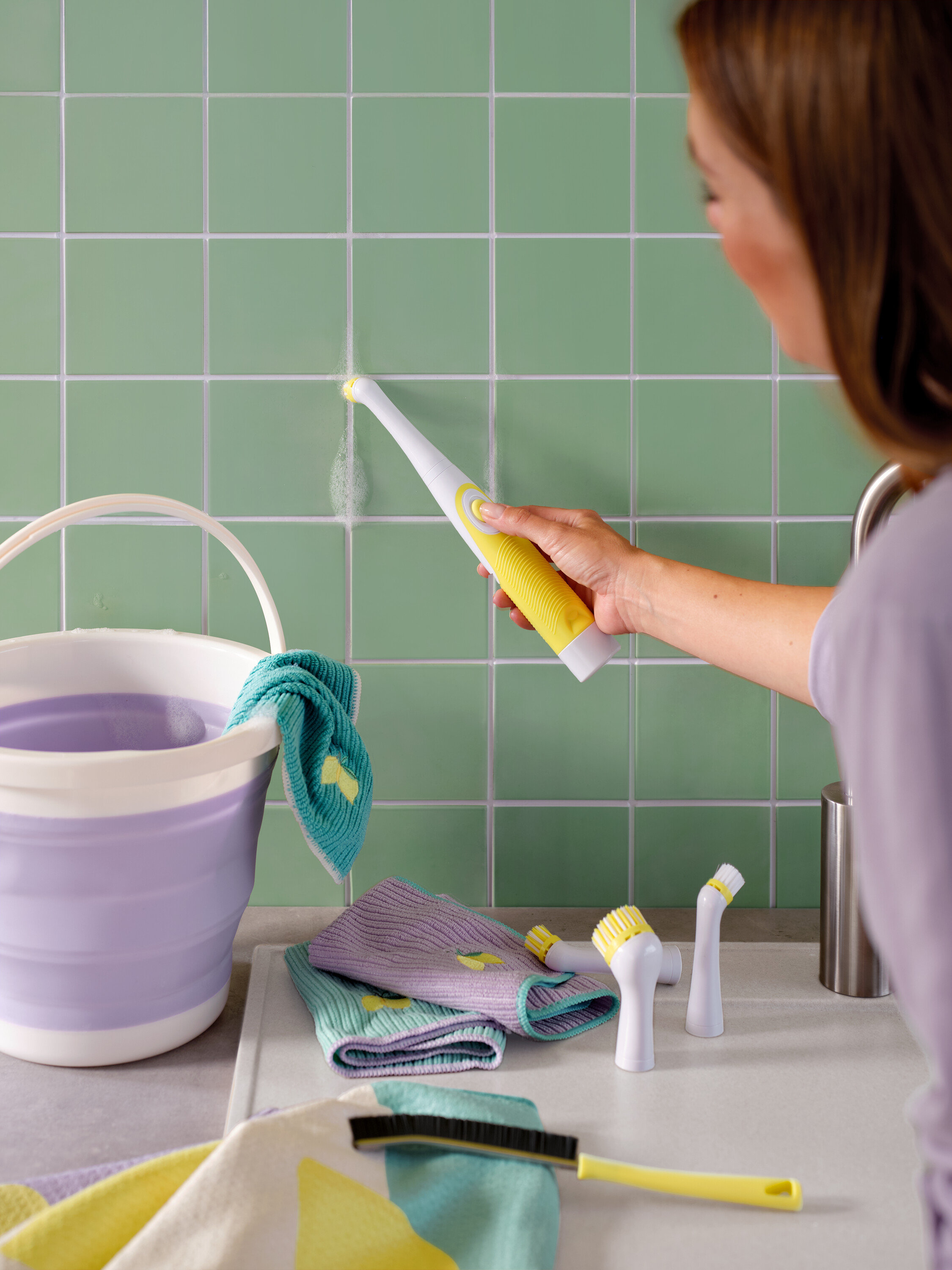 Frau reinigt grüne Fliesen mit einer gelb-weissen Bürste, Eimer und Tücher sind ebenfalls sichtbar.