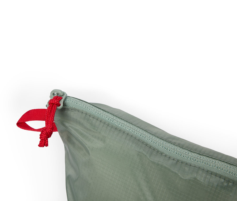 Nahaufnahme einer olivgrünen Tasche mit Reißverschluss und roter Zugschnur vor weißem Hintergrund.