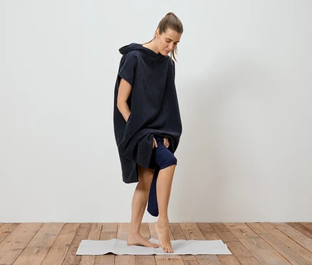 Frau im marineblauen Badeponcho, der bis zu den Oberschenkeln reicht, steht barfuß auf einer Yogamatte.