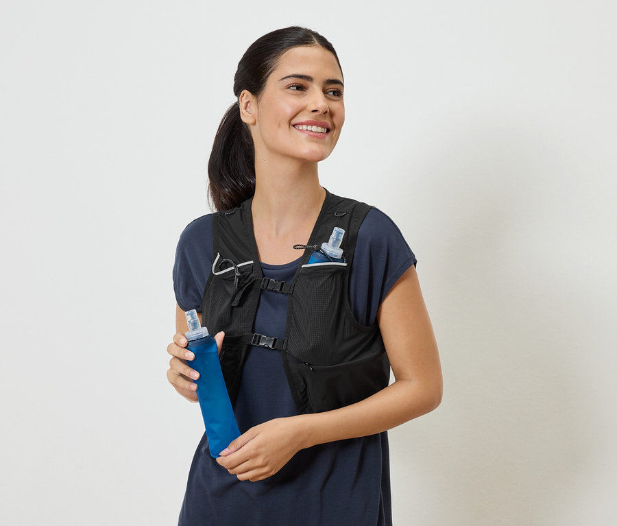 Lächelnde Frau mit schwarzer Laufweste und blauer Trinkflasche in der Hand.