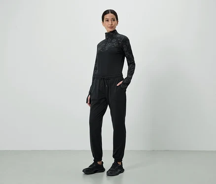 Frau in schwarzem, gemustertem Oberteil und Jogginghose mit Händen in den Taschen, dazu schwarze Turnschuhe.