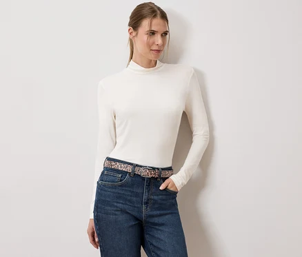 Frau mit cremefarbenem Rollkragenpullover, Jeans und Gürtel mit Tiermuster steht vor einer weißen Wand.