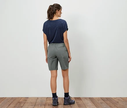 Rückansicht einer Frau mit dunklem T-Shirt, grauen Shorts und Wanderschuhen vor einer weißen Wand.