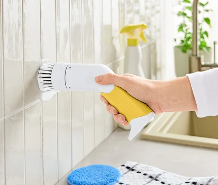 Eine Hand hält eine elektrische Reinigungsbürste mit gelbem Griff und weißem Bürstenkopf, um Fliesen zu reinigen.