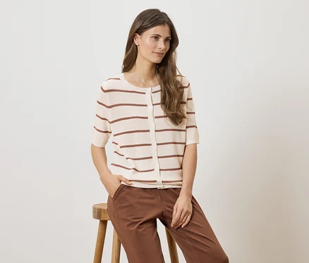 Frau sitzt auf einem Hocker, trägt eine gestreifte, kurzärmelige Strickjacke und braune Hose.