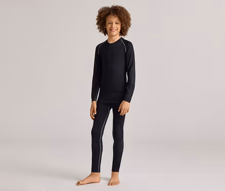Ein lächelnder kleiner Junge mit lockigem Haar steht barfuß und trägt ein schwarzes Langarmshirt und passende schwarze Leggings mit hellgrauen Nähten.