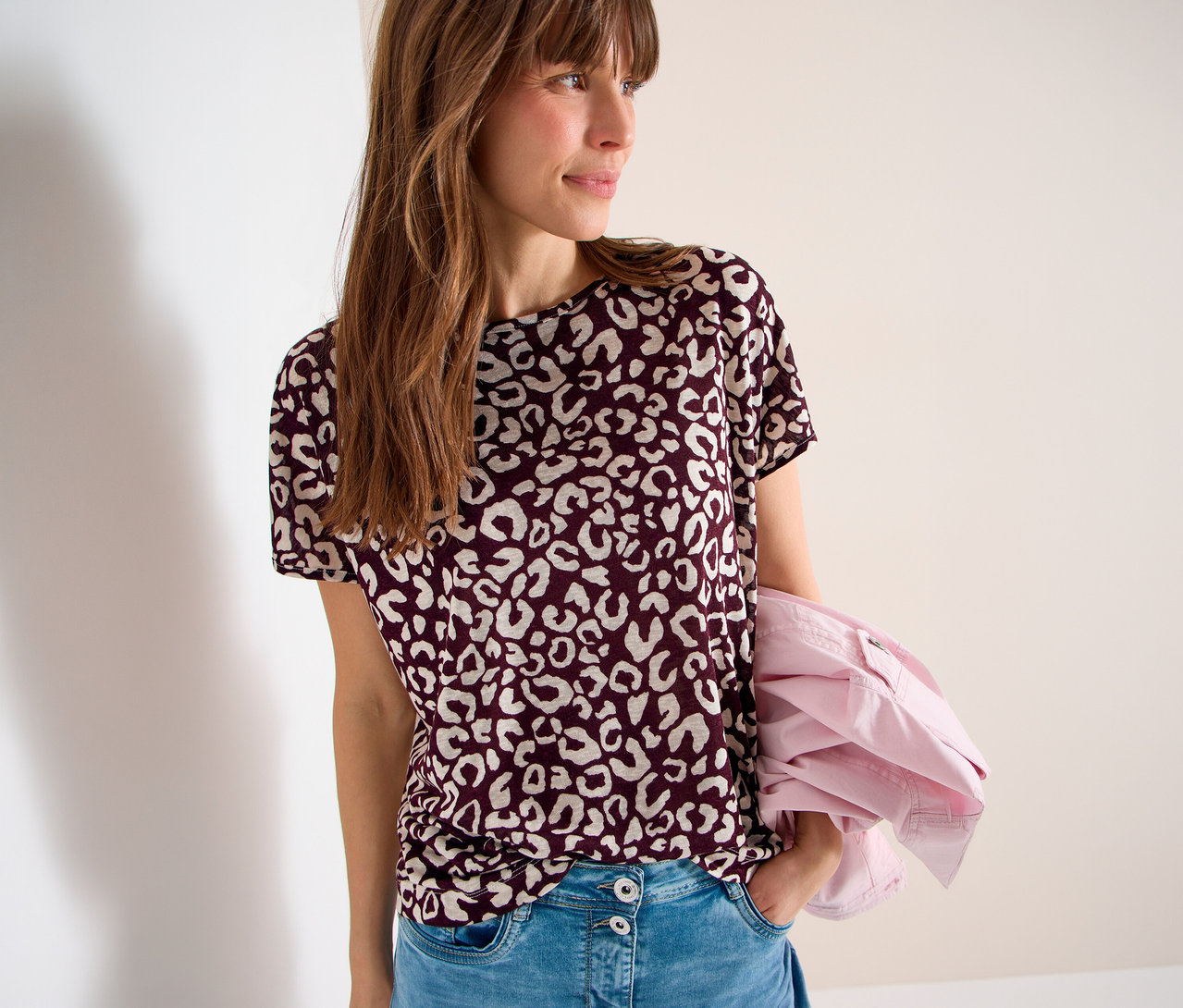 Frau mit burgunderfarbenem T-Shirt mit Leopardenmuster und Jeans, eine rosa Jacke über dem Arm.