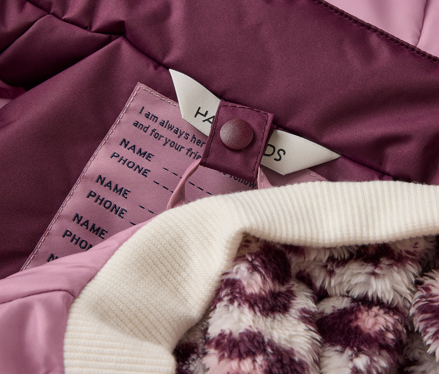 Nahaufnahme des Innenfutters einer lila Jacke mit einem weißen, gerippten Bündchen und einem flauschigen, gemusterten Futter. Ein Etikett mit Feldern für Name und Telefonnummer ist sichtbar.