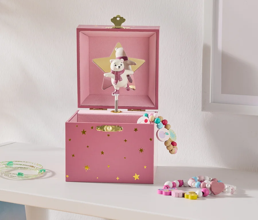Eine offene rosa Spieluhr mit goldenen Sternen und einem tanzenden Eisbären auf einem goldenen Stern, daneben bunte Perlenarmbänder auf einem weißen Regal.
