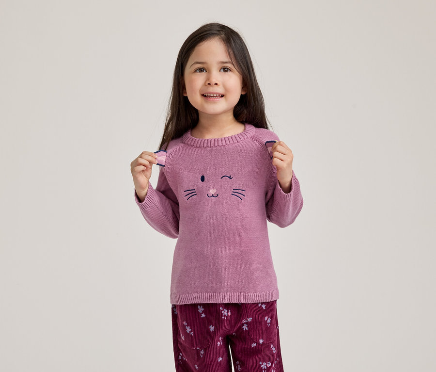 Ein lächelndes kleines Mädchen mit dunklen Haaren trägt einen rosa Pullover mit einem zwinkernden Katzengesicht und eine dunkelrote Cordhose mit Blumenmuster. Sie hält die kleinen Ohren-Details an den Schultern ihres Pullovers hoch.
