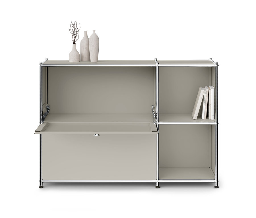 Hellgrauer Modulschrank mit Chromrahmen, links mit einer Klapptür, die einen Schreibtisch bildet, und rechts mit zwei offenen Fächern. Oben stehen drei Dekovasen, im oberen rechten Fach Bücher.