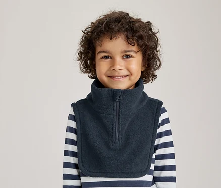 Lächelndes Kleinkind mit lockigem Haar, das einen dunkelblauen Fleece-Halswärmer über einem gestreiften Langarmshirt trägt.