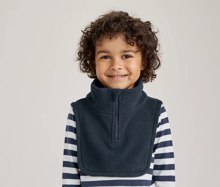 Lächelndes Kleinkind mit lockigem Haar, das einen dunkelblauen Fleece-Halswärmer über einem gestreiften Langarmshirt trägt.