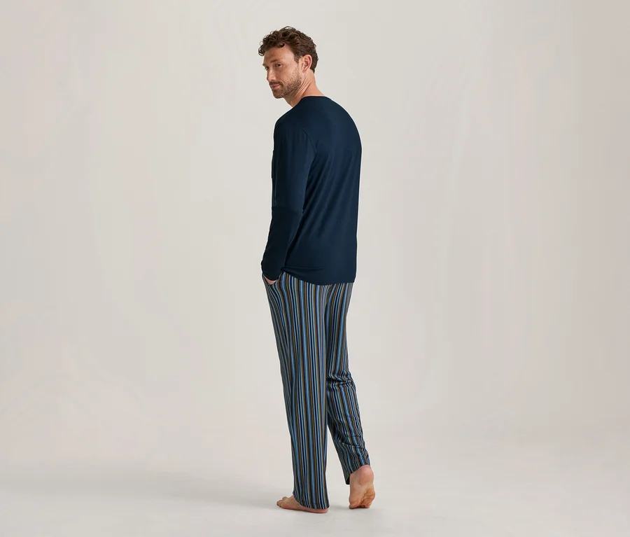 Mann von hinten, über die linke Schulter blickend, in marineblauem Langarmshirt und blau-braun-hellblau gestreifter Pyjamahose, barfuß stehend.