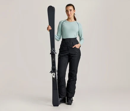 Eine Frau steht, hält einen schwarzen Ski aufrecht und trägt ein hellgrünes Langarmshirt sowie eine schwarze, hoch taillierte Skihose.