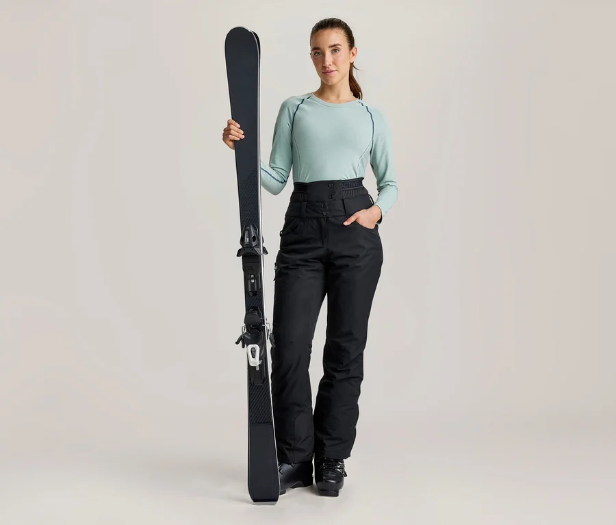Eine Frau steht, hält einen schwarzen Ski aufrecht und trägt ein hellgrünes Langarmshirt sowie eine schwarze, hoch taillierte Skihose.