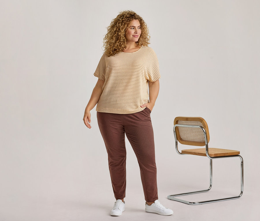 Eine Frau mit lockigem blondem Haar, die ein hell gestreiftes Oberteil, eine dunkelbraune Hose und weiße Turnschuhe trägt, steht neben einem modernen Holzstuhl.