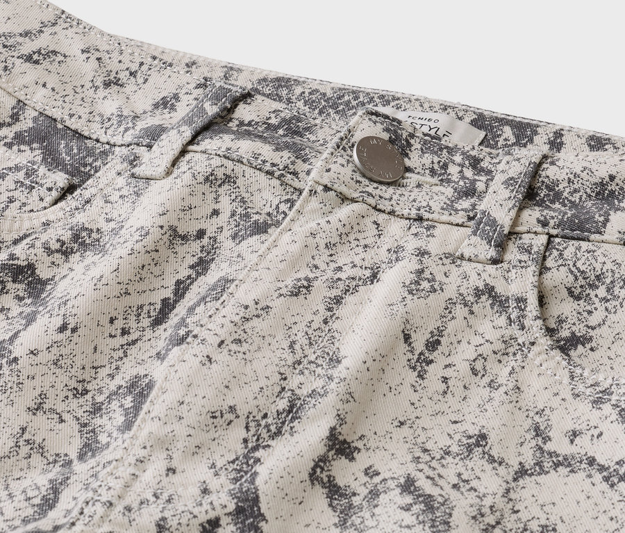 Nahaufnahme einer hellbeigen Hose mit dunkelgrauem, meliertem Muster, die einen silbernen Knopf, Gürtelschlaufen und ein „TCHIBO STYLE“-Etikett zeigt.