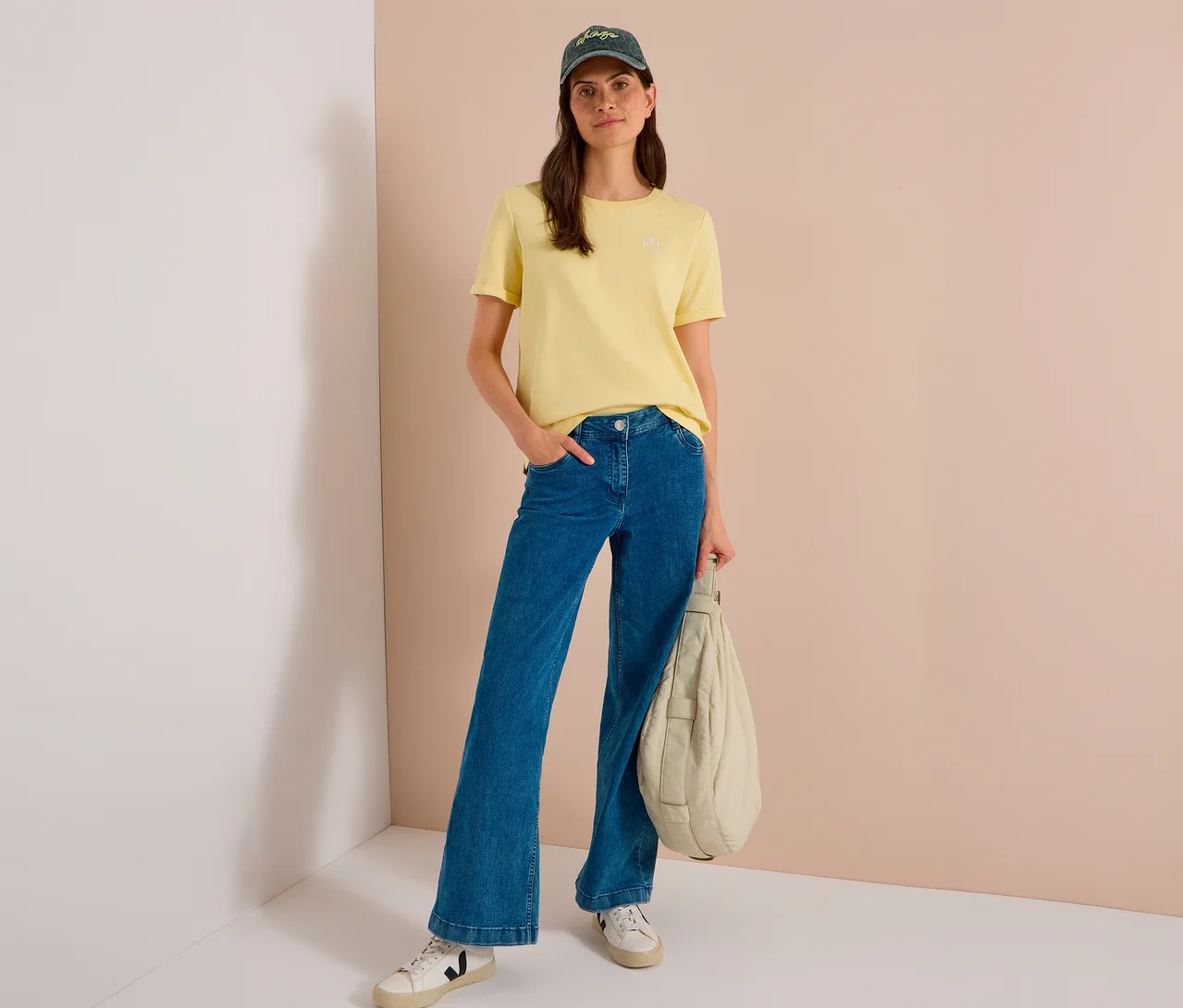 Frau trägt gelbes T-Shirt, blaue weite Jeans, weiße Sneaker und eine dunkelgrüne CECIL Baseballcap, hält eine hellfarbene, gepolsterte Tasche.