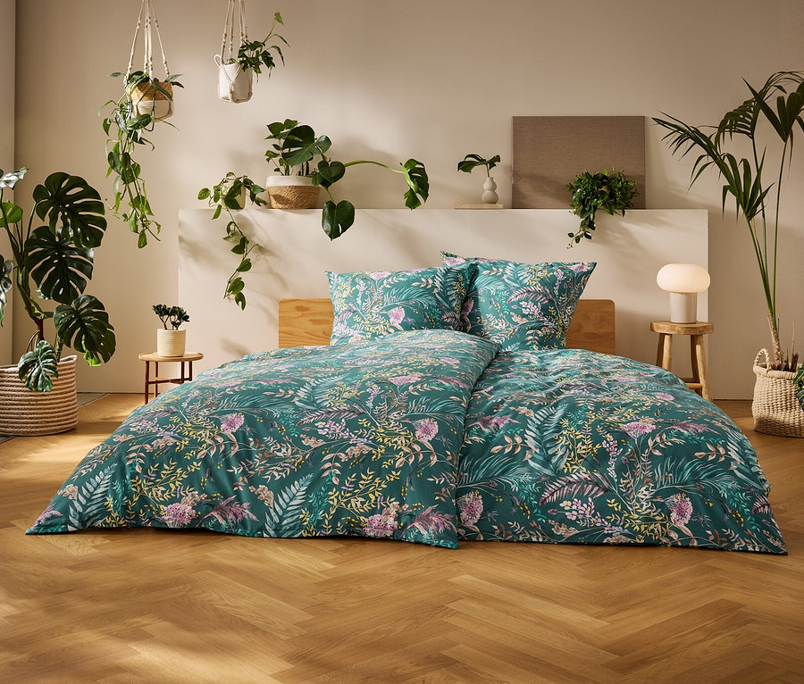 Ein Bett mit grüner Renforcé-Bettwäsche mit Blumenmuster in einem hellen Schlafzimmer mit Holzfußboden und vielen Topfpflanzen.