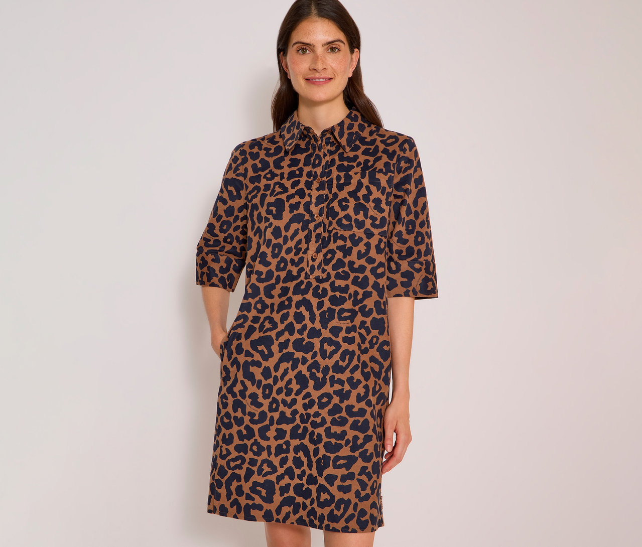 Lächelnde Frau in einem braun-dunkelblauen CECIL Hemdblusenkleid mit Leopardenmuster und Dreiviertelärmeln, Hände in den Taschen.