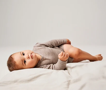 Baby liegt auf dem Rücken auf einem hellen Laken, trägt einen gerippten, langärmeligen, taupefarbenen Body und blickt in die Kamera.