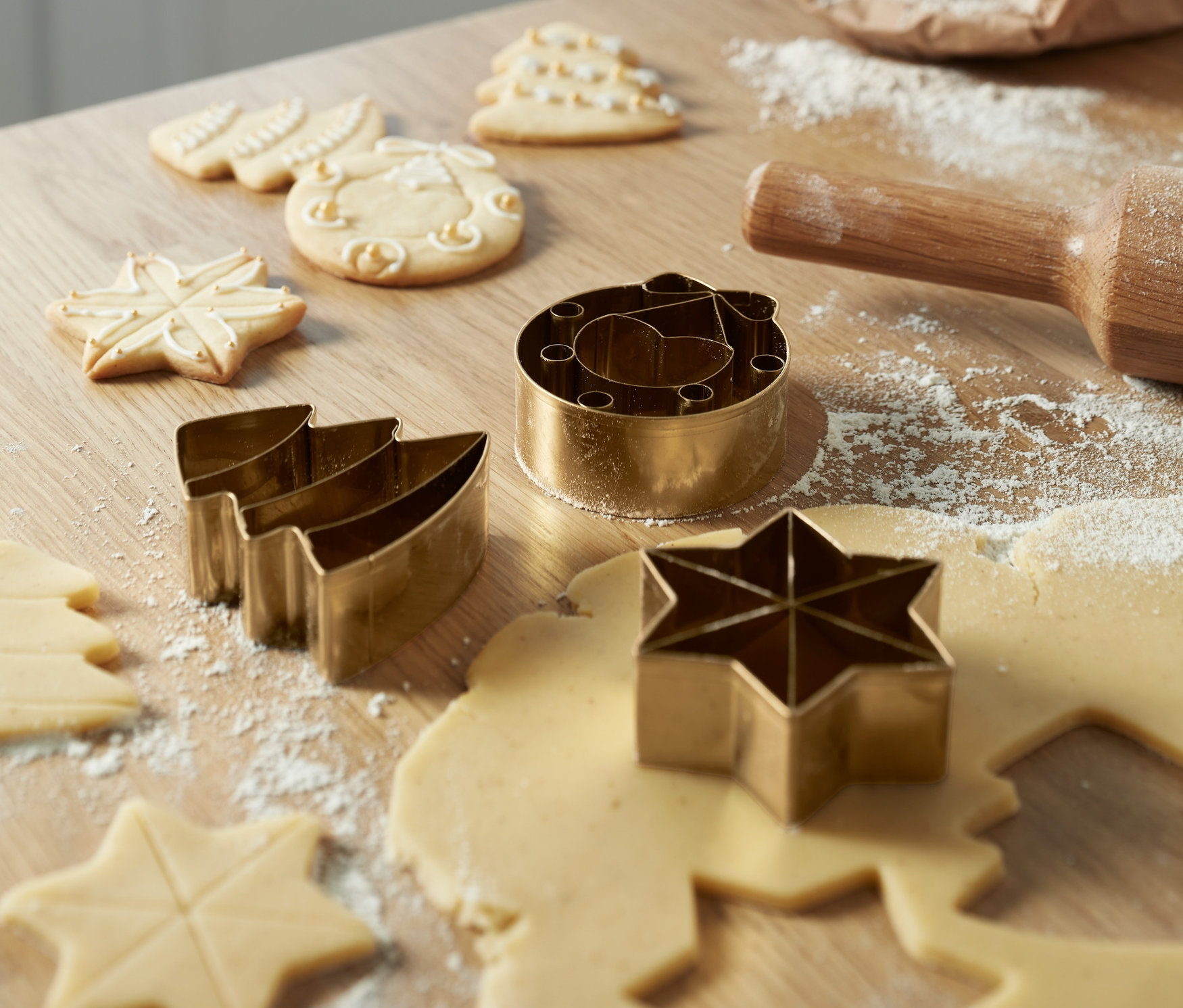 Auf einem Holztisch liegen Premium-Keksausstecher-Sets in Form von Sternen und Weihnachtsbäumen, zusammen mit Teig, einem Nudelholz und einem Sack Mehl.