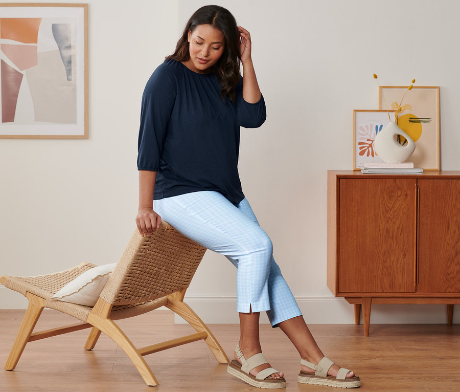 Eine Frau sitzt auf einem Stuhl und trägt ein dunkelblaues Shirt mit 3/4-Arm, eine hellblau-weiße 7/8-Stretchhose und Plateau-Sandale aus Leder.