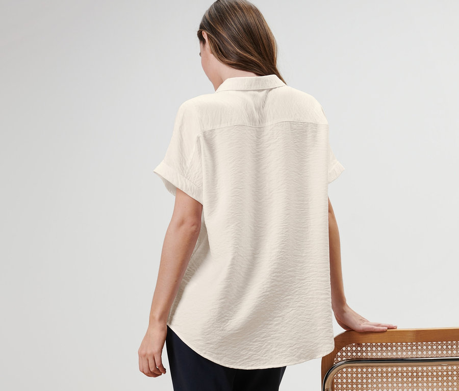 Frau steht mit dem Rücken zur Kamera und trägt eine Bluse, während sie sich an einen Stuhl lehnt.