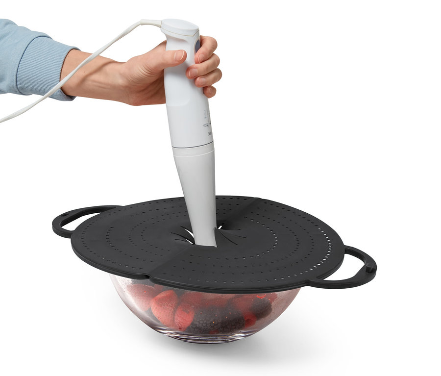 Eine Hand hält einen Stabmixer durch einen schwarzen Pfannenspritzschutz über einer Glasschüssel mit Früchten.
