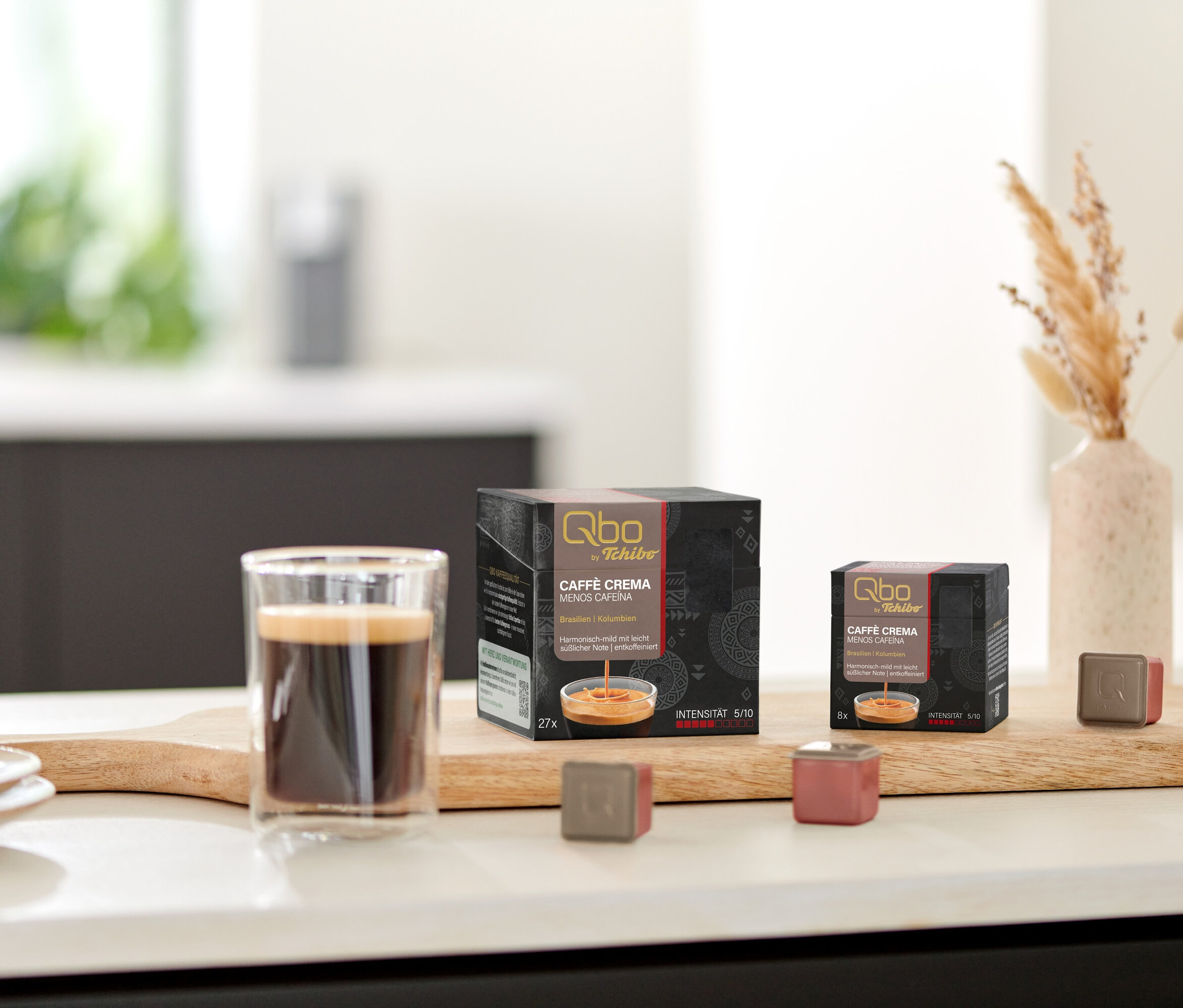 Auf einem Tisch stehen Qbo Caffè MENOS CAFEÍNA Kapselpackungen und eine Tasse Kaffee auf einem Holzbrett.