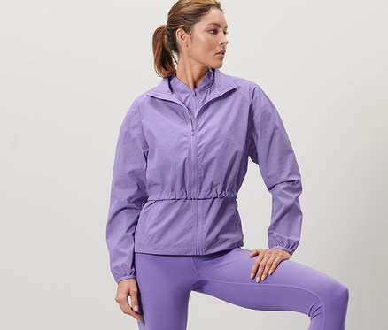 Frau posiert in lila 3-in-1-Laufjacke und Leggings, ein Bein auf einer Holzkiste.