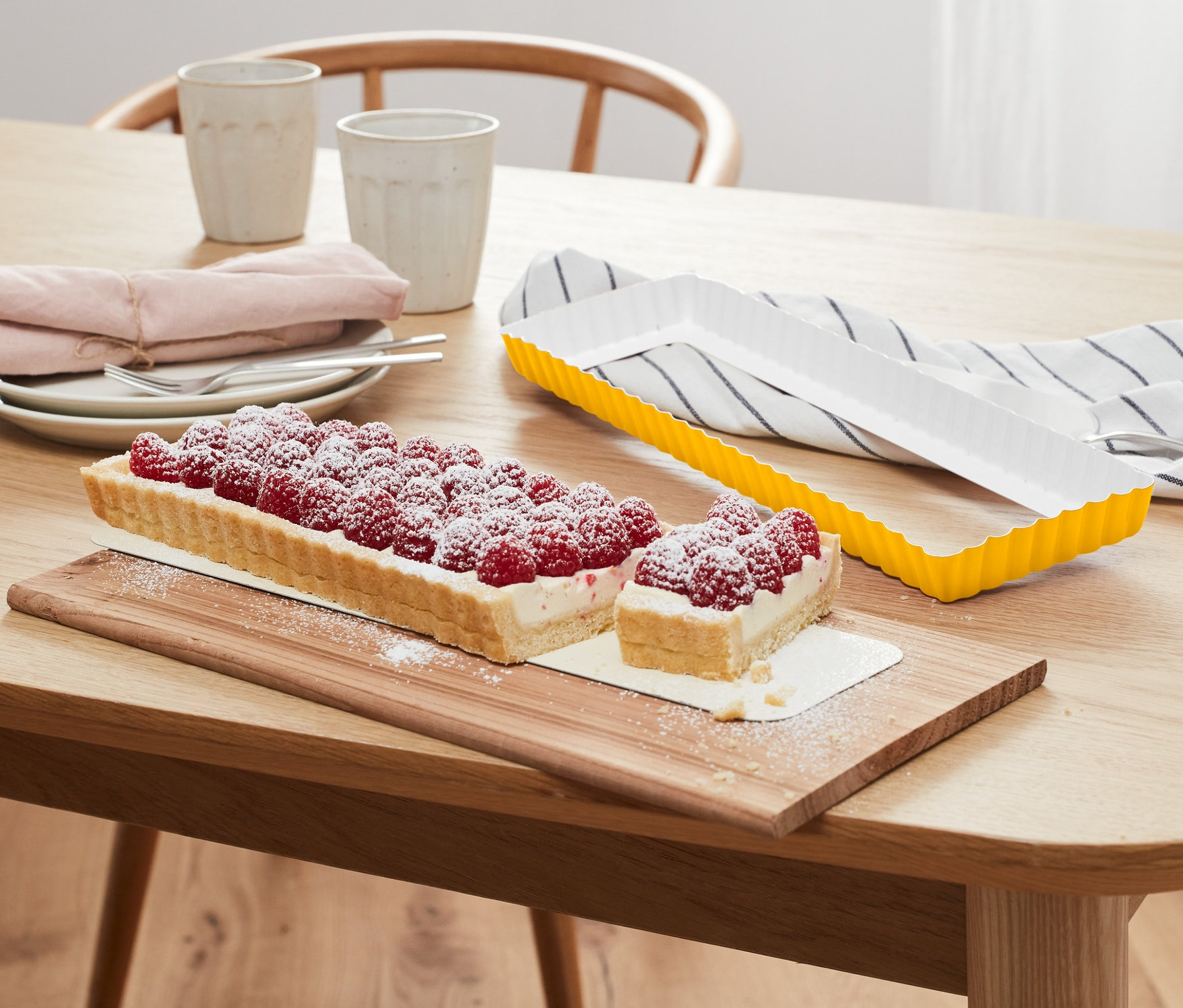 Rechteckige Himbeertarte auf Holzbrett neben gelber Tarteform.