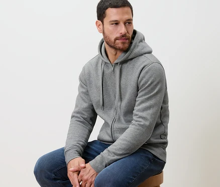 Mann sitzt auf einem Hocker und trägt eine graue Kapuzenjacke.