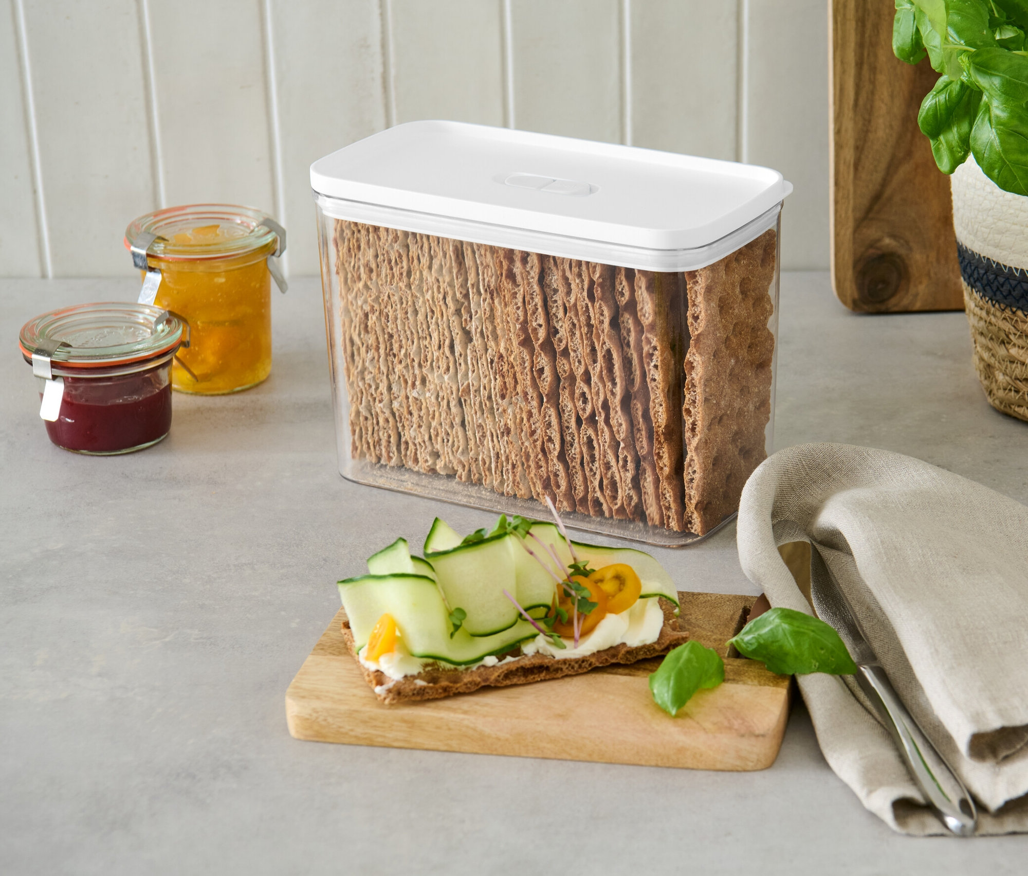Ein Vorratsbehälter mit weißem Deckel, gefüllt mit Brotcrackern, steht auf einer Theke neben Marmeladengläsern.