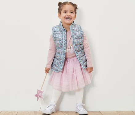 Lächelndes Mädchen trägt eine geblümte Kinder-Steppweste, ein rosa Langarmshirt, einen rosa Rock und weiße Turnschuhe und hält einen Zauberstab.