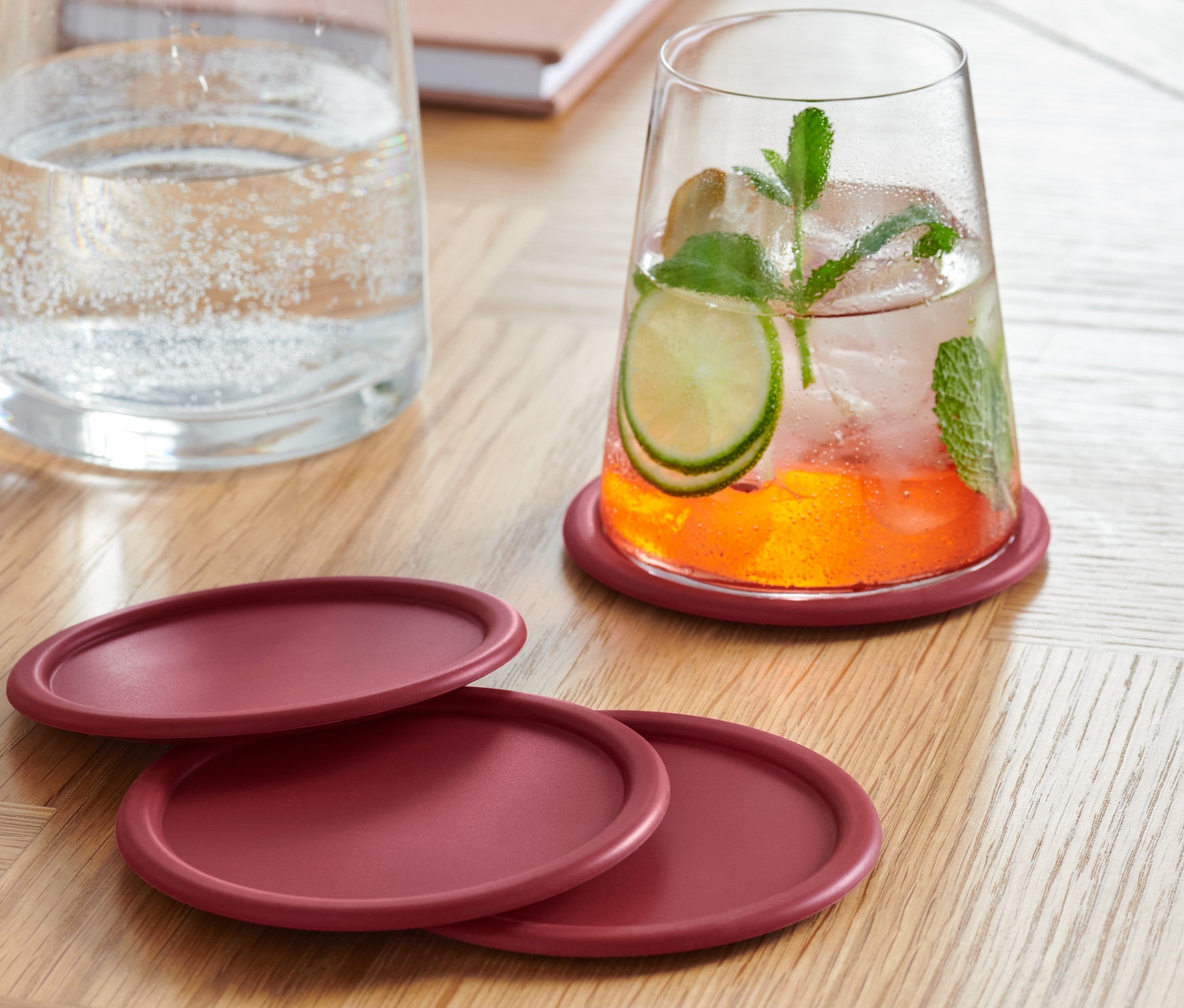 Ein mit einem Getränk gefülltes Glas steht auf einem der vier bordeauxroten Glasuntersetzer. Drei weitere Untersetzer liegen auf einem Holztisch. Im Hintergrund stehen eine Wasserkaraffe und ein Notizbuch.